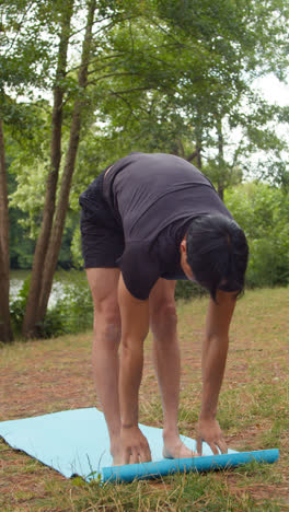 Vertical-Video-Of-Man-Wearing-Sports-Clothing-Doing-Yoga-Stretches-On-Mat-In-Forest-By-Lake-Or-River-Enjoying-Peace-And-Beauty-Of-Nature-Shot-In-Real-Time-3